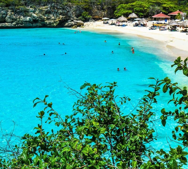 A hidden Caribbean beach with crystal-clear turquoise water, swimmers in the bay, and a quiet white-sand shoreline lined with thatched umbrellas and sunbeds, surrounded by lush green foliage and rocky cliffs.