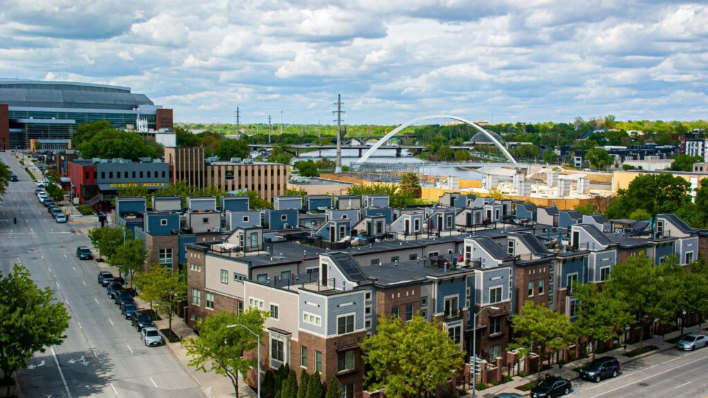 A vibrant cityscape in Iowa featuring rows of modern townhomes, tree-lined streets, and a distinctive arched bridge spanning a river under a partly cloudy sky.