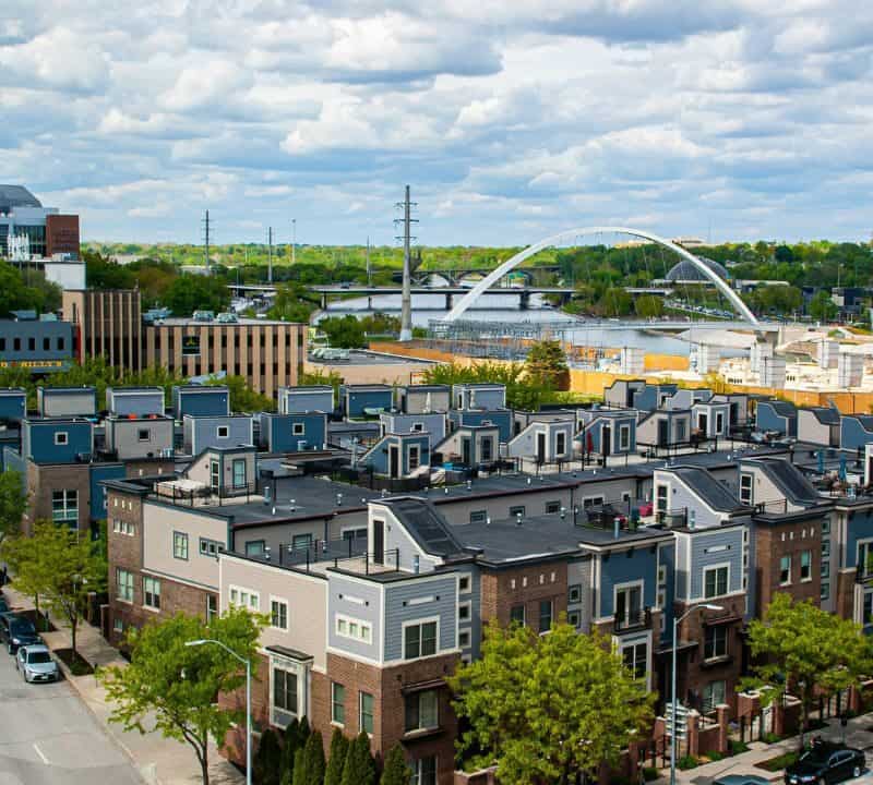 A vibrant cityscape in Iowa featuring rows of modern townhomes, tree-lined streets, and a distinctive arched bridge spanning a river under a partly cloudy sky.
