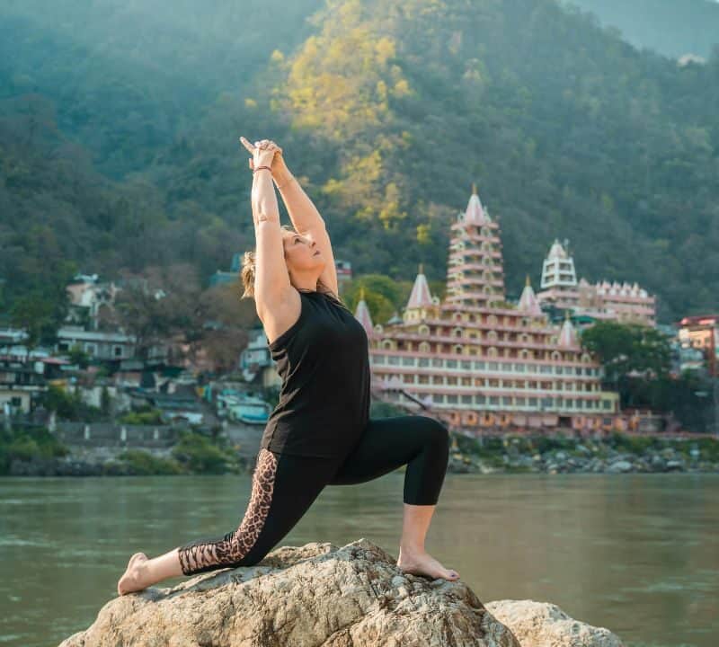 Woman practicing a yoga pose on a rock beside the Ganges River, with the iconic pink and white Tera Manzil Temple and forested hills of Rishikesh in the background.
