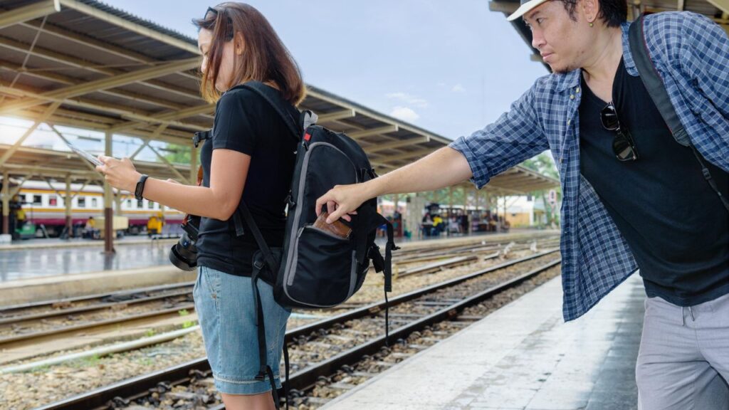 Man stealthily stealing a wallet from a woman’s backpack at a train station while she is distracted by her phone.