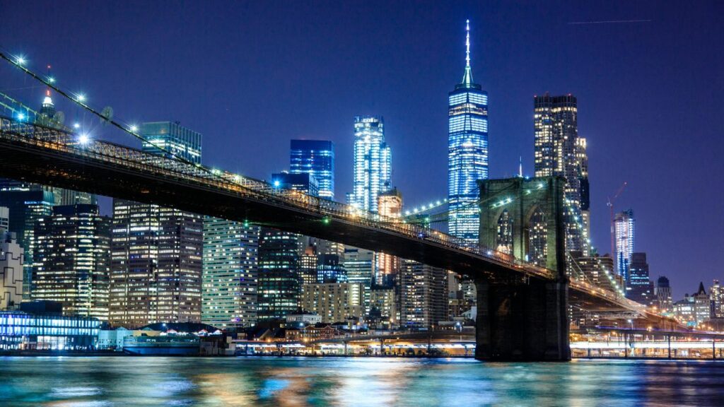 The Brooklyn Bridge stretches across the East River at night with the illuminated Manhattan skyline and One World Trade Center glowing in the background.