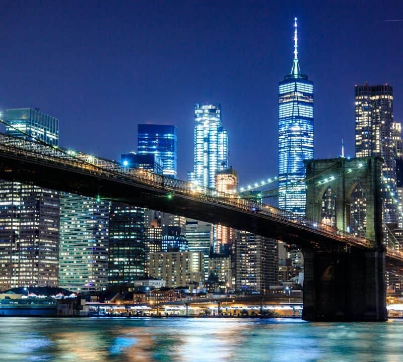 The Brooklyn Bridge stretches across the East River at night with the illuminated Manhattan skyline and One World Trade Center glowing in the background.