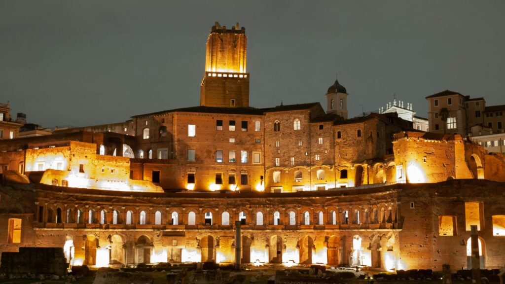 Trajan's Market in Rome illuminated at night, showcasing ancient arches, windows, and the tall Torre delle Milizie.