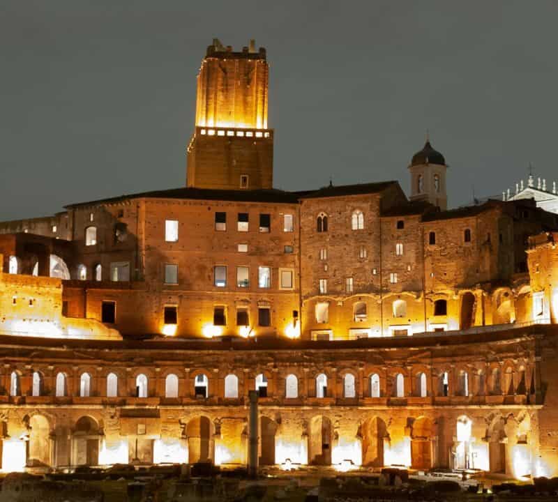 Trajan's Market in Rome illuminated at night, showcasing ancient arches, windows, and the tall Torre delle Milizie.