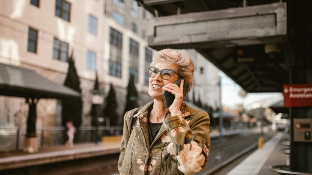Smiling senior woman talking on the phone at a train station, representing joyful and budget-friendly retirement travel.