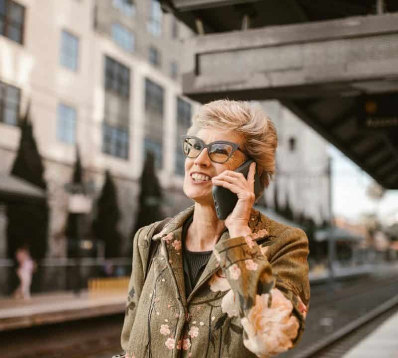 Smiling senior woman talking on the phone at a train station, representing joyful and budget-friendly retirement travel.