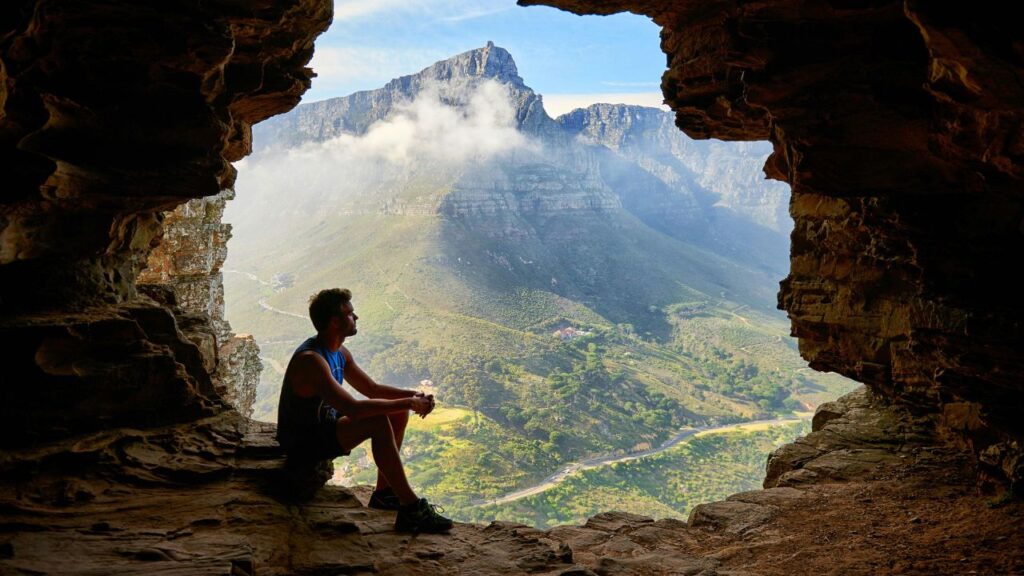 A man sits at the edge of a rocky cave opening, looking out over a scenic mountain valley with winding roads and mist-covered peaks.