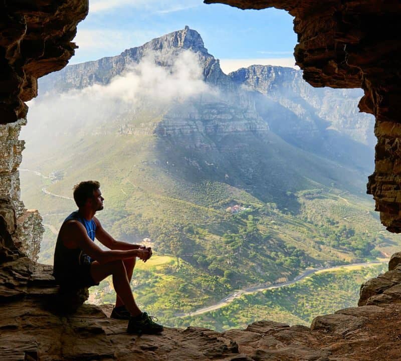 A man sits at the edge of a rocky cave opening, looking out over a scenic mountain valley with winding roads and mist-covered peaks.