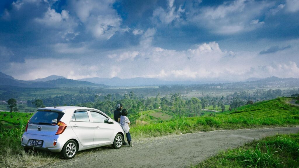 Couple leans on a white car parked on a rural road, gazing at a wide valley and green hills under a dramatic sky.