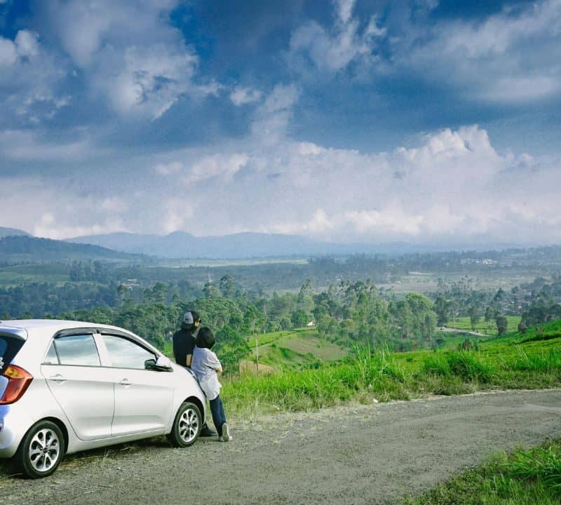 Couple leans on a white car parked on a rural road, gazing at a wide valley and green hills under a dramatic sky.