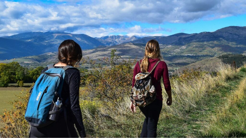Two women with backpacks hiking through grassy hills, facing scenic mountain views under a partly cloudy sky.