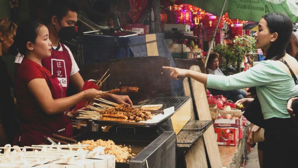 A busy street food stall with skewers grilling on an open flame, as a vendor serves a customer in a lively outdoor market.