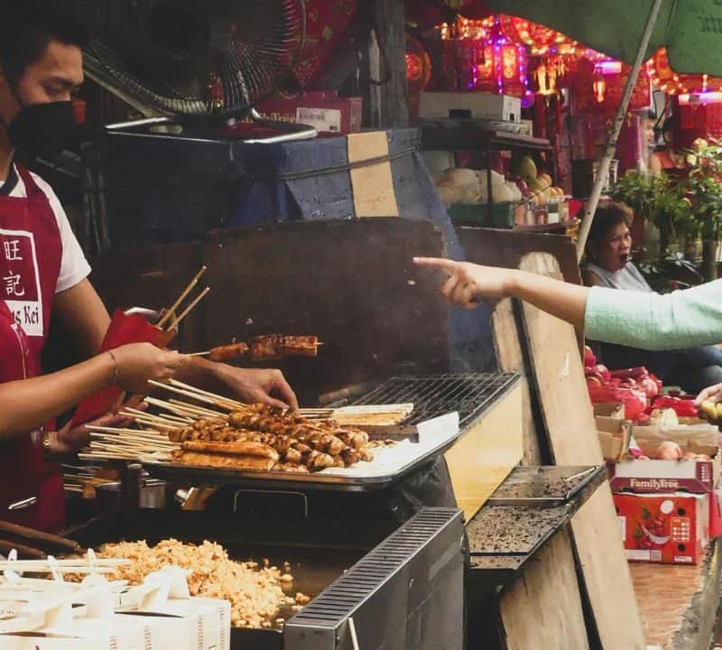 A busy street food stall with skewers grilling on an open flame, as a vendor serves a customer in a lively outdoor market.