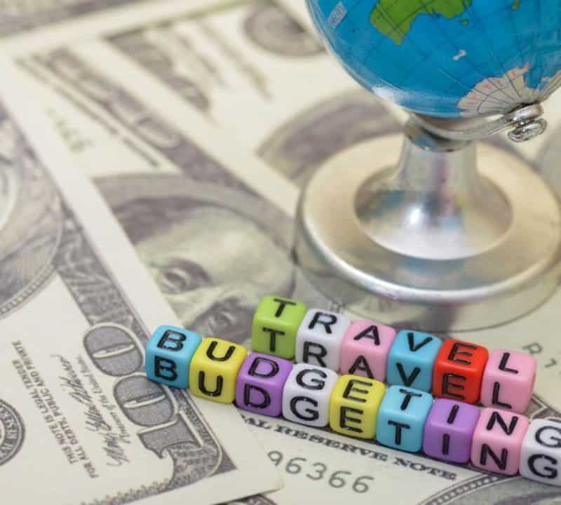 Colorful alphabet beads spelling "Travel Budgeting" placed on U.S. dollar bills with a small globe in the background