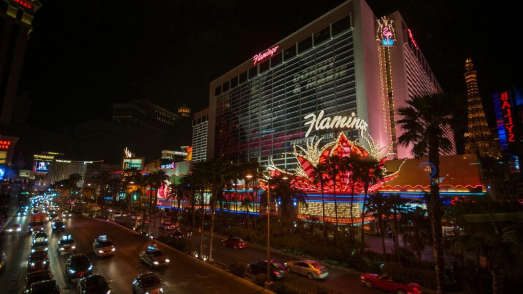 View of the Flamingo Hotel and Casino on the Las Vegas Strip at night, with bright neon lights, busy streets filled with cars, and other iconic signs like the Eiffel Tower replica in the background.