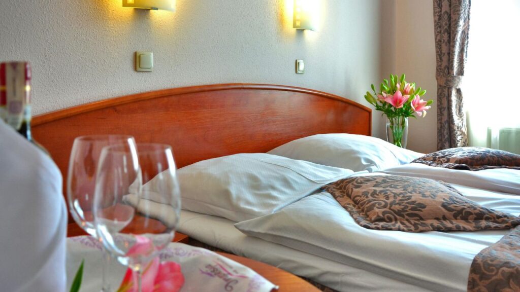 Neatly made hotel bed with decorative pillows, wine glasses, and a vase of fresh pink lilies near a window.