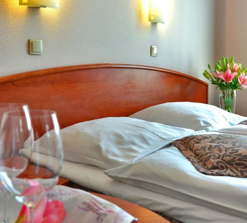 Neatly made hotel bed with decorative pillows, wine glasses, and a vase of fresh pink lilies near a window.