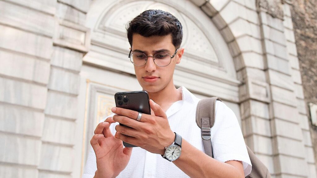 Young man wearing glasses and a backpack looking at his smartphone while standing in front of a stone building.