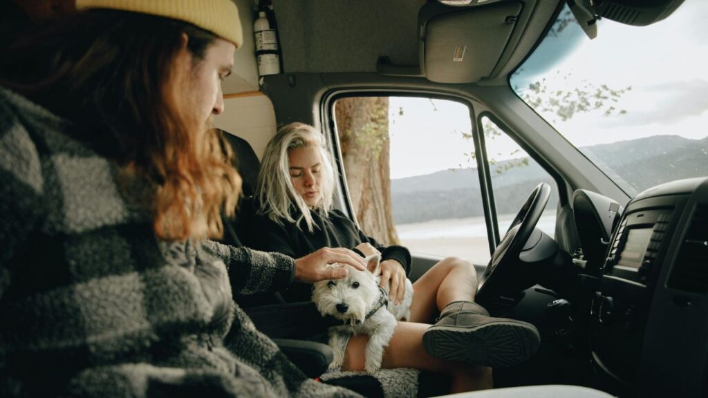 A couple relaxes inside a camper van with their small white dog during a scenic road trip by a lake or forest.