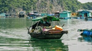 Vendor boat selling snacks and fruits on calm water, with floating houses and cliffs in the background
