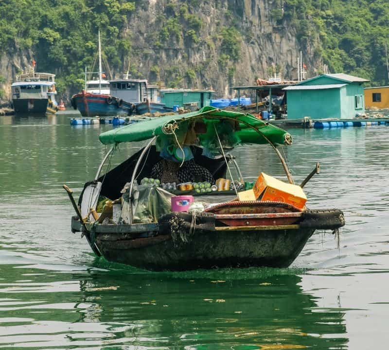 Vendor boat selling snacks and fruits on calm water, with floating houses and cliffs in the background