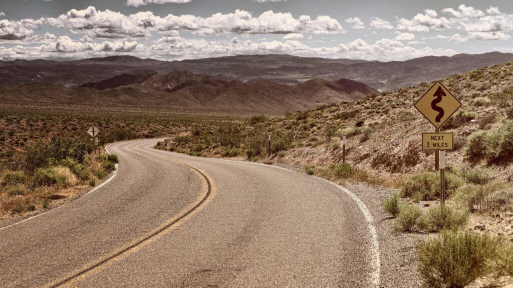 A winding desert road with a caution sign reading "Next 3 Miles," surrounded by dry hills and mountains under a partly cloudy sky.