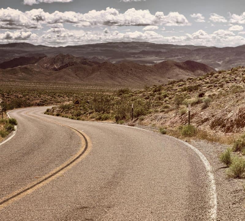 A winding desert road with a caution sign reading "Next 3 Miles," surrounded by dry hills and mountains under a partly cloudy sky.