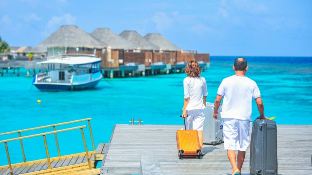 Couple walking with luggage toward overwater bungalows in a tropical resort, symbolizing luxury travel and hidden costs.