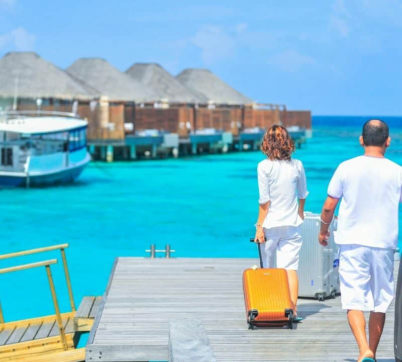 Couple walking with luggage toward overwater bungalows in a tropical resort, symbolizing luxury travel and hidden costs.