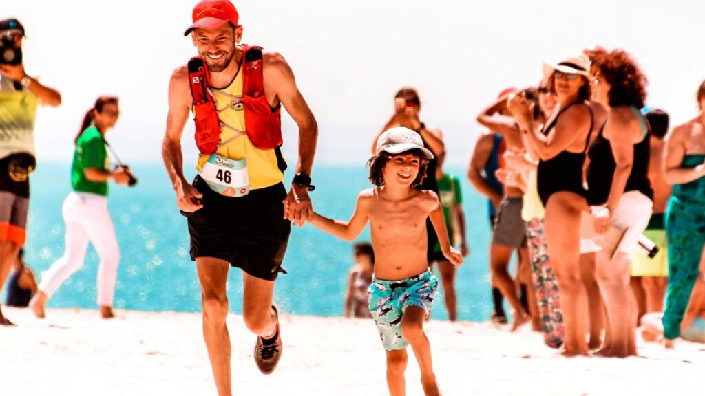 Smiling father and son running on the beach during a family race event, surrounded by cheering people on a sunny day.