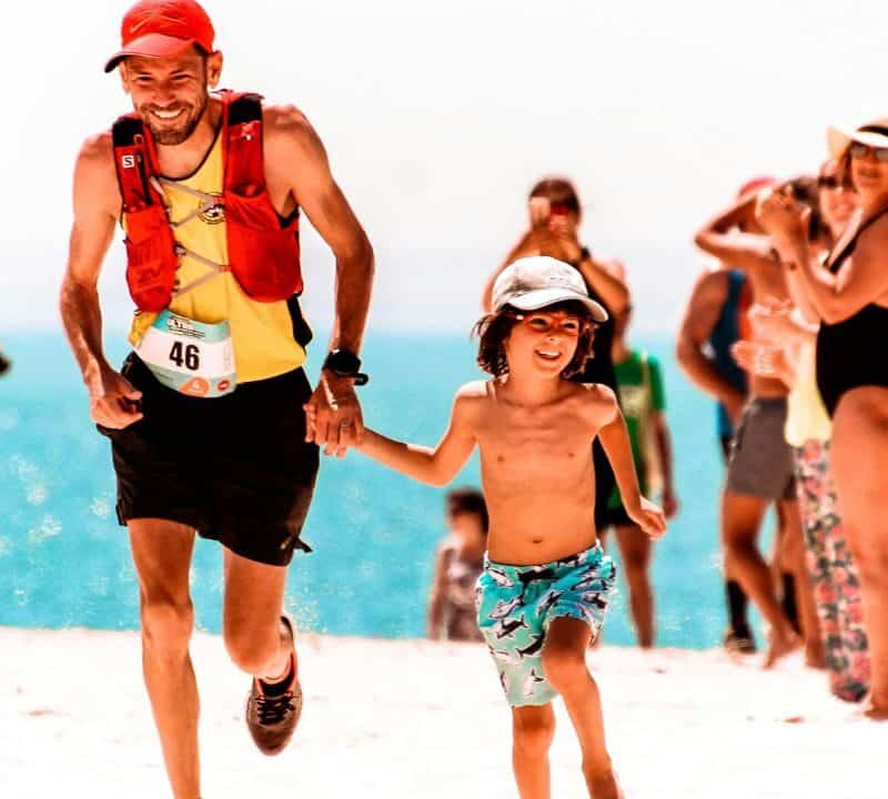 Smiling father and son running on the beach during a family race event, surrounded by cheering people on a sunny day.