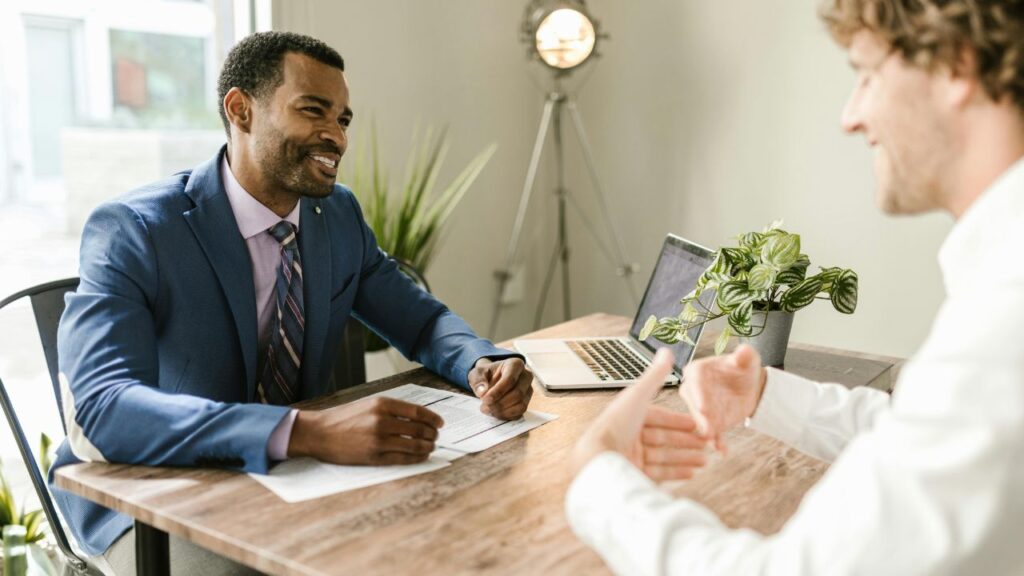Smiling insurance advisor in blue suit reviews paperwork with client across desk, laptop and potted plant nearby.