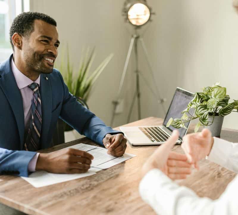 Smiling insurance advisor in blue suit reviews paperwork with client across desk, laptop and potted plant nearby.