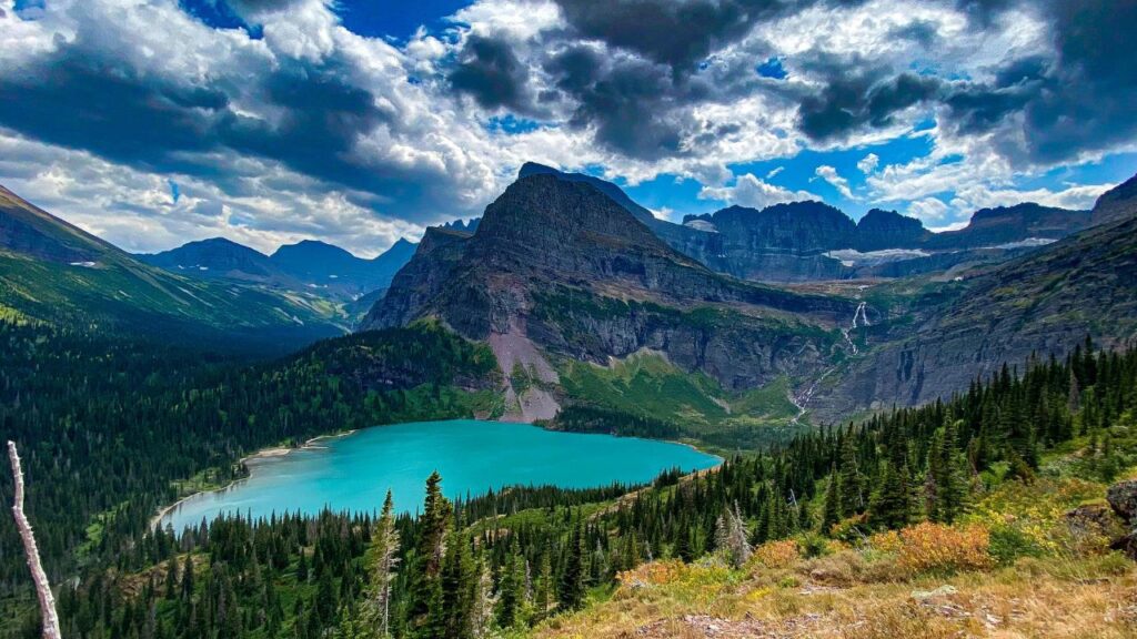Turquoise alpine lake surrounded by dense forest and towering mountain peaks under dramatic clouds in a scenic wilderness landscape.