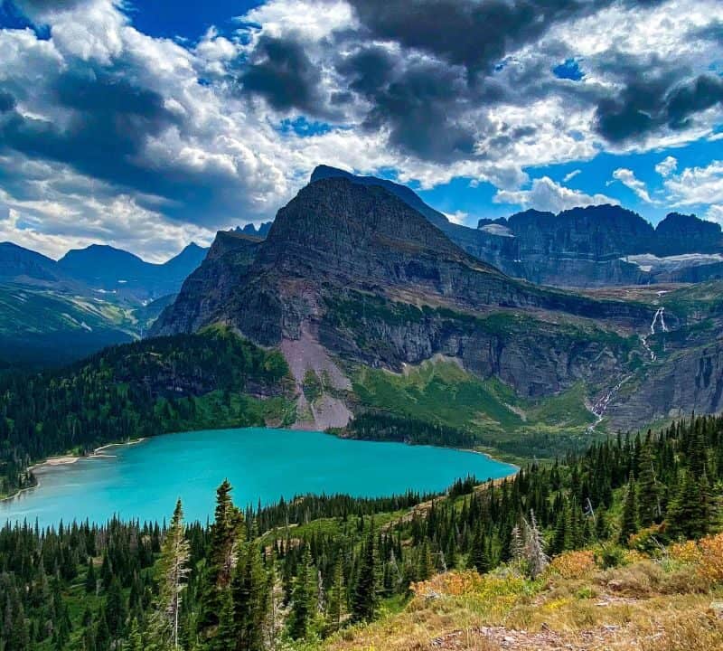 Turquoise alpine lake surrounded by dense forest and towering mountain peaks under dramatic clouds in a scenic wilderness landscape.