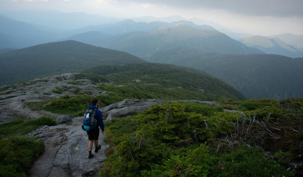 A solo hiker with a backpack walks along a rocky ridge in the Adirondack Mountains, surrounded by misty green peaks.