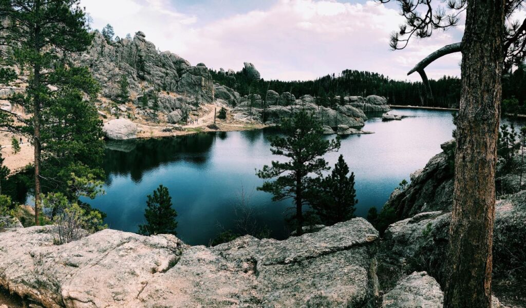 Rocky cliffs and pine trees surround a calm mountain lake under a partly cloudy sky in a rugged forested landscape.