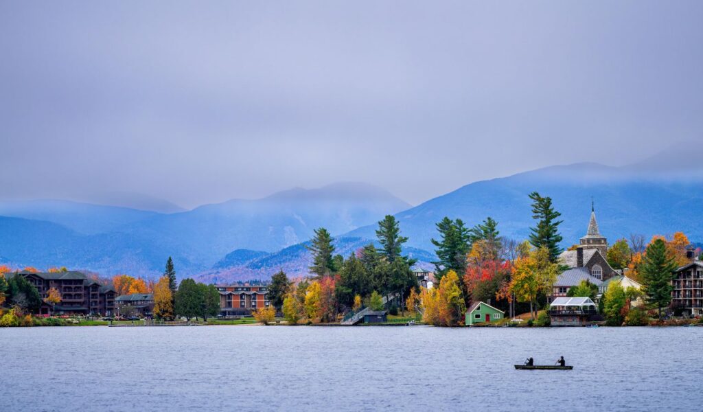 Canoe with two people on a calm lake, surrounded by colorful autumn trees, cozy buildings, and misty Adirondack mountains.