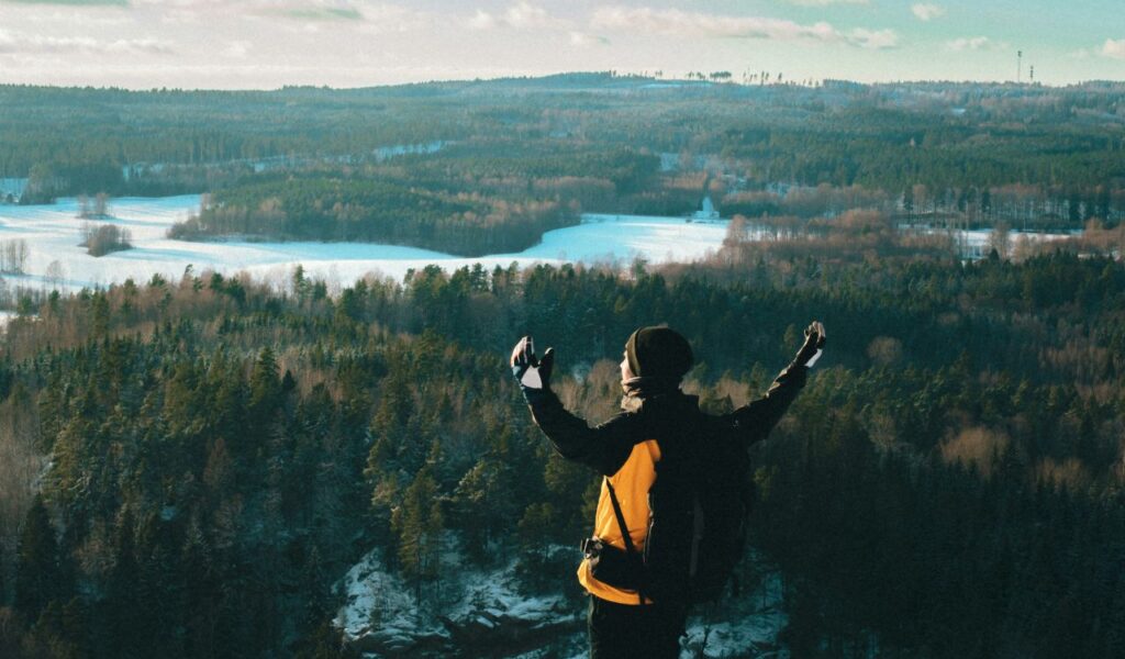A hiker with arms raised stands on a high cliff overlooking a vast winter forest and snow-covered landscape.