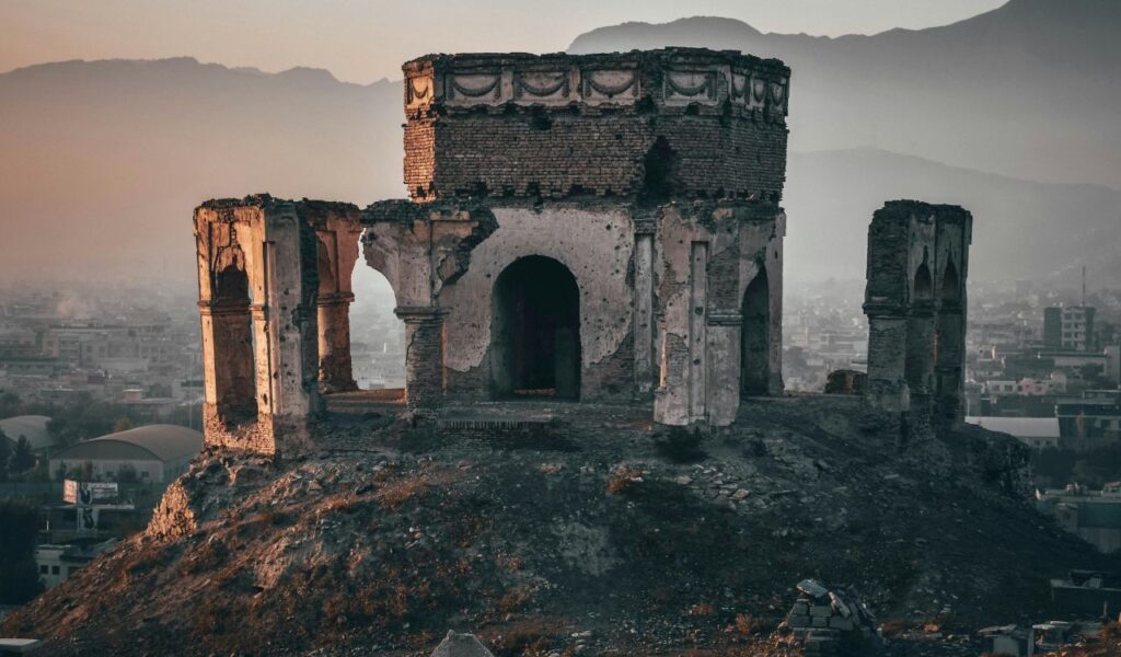 A crumbling historic fortress stands on a hilltop overlooking a hazy cityscape with mountains in the background.