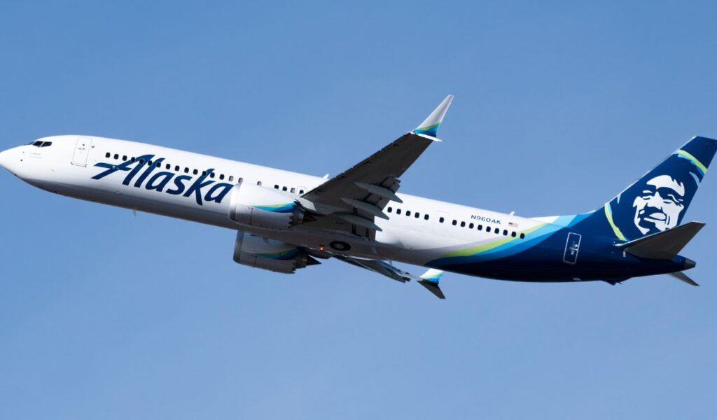 Alaska Airlines plane soars through a clear blue sky, featuring its signature Eskimo logo on the tail.