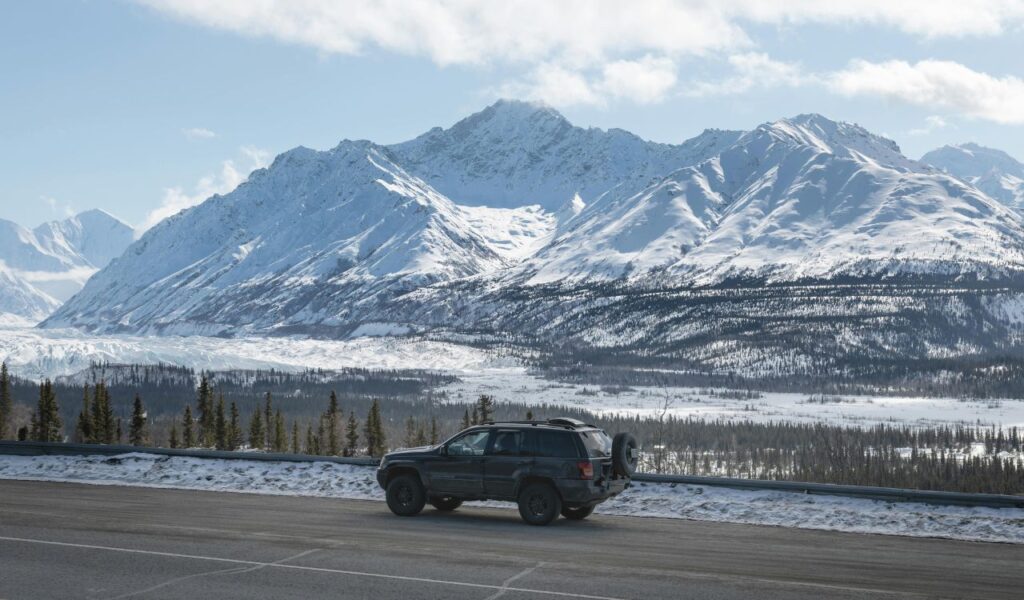 A black SUV parked on a snowy roadside overlooks a vast Alaskan valley with towering snow-covered mountains in view.