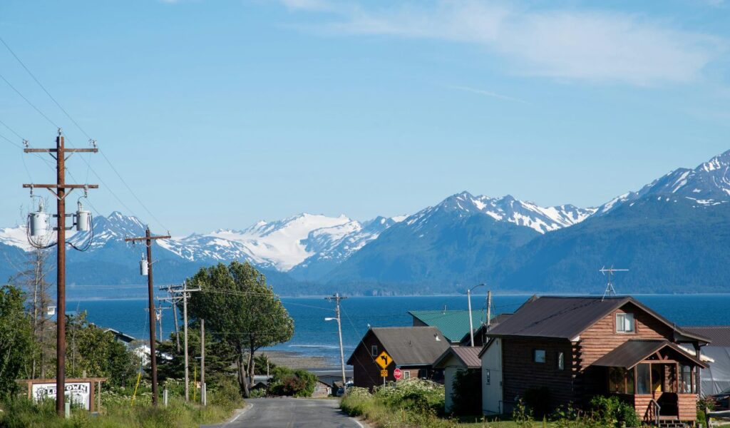 A quiet coastal road leads to the sea, with wooden cabins on one side and snow-capped Alaskan mountains rising across the bay under a clear blue sky.