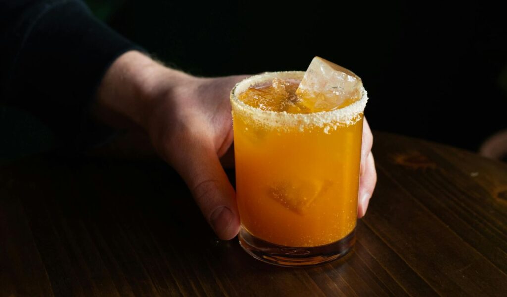 Hand holding an alcoholic cocktail with orange liquid, ice, and a salted rim, placed on a wooden surface.