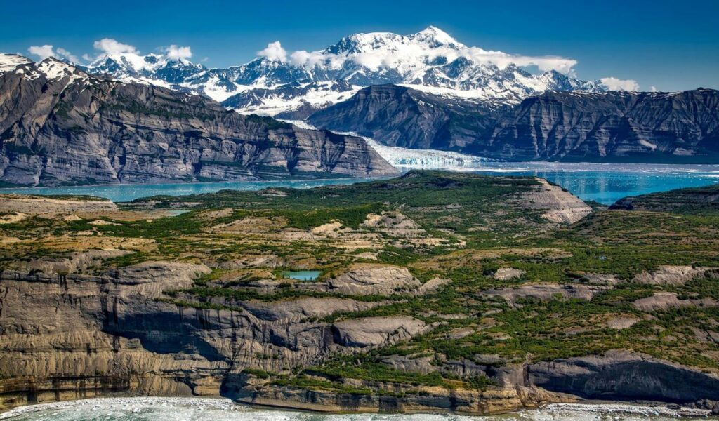 Snow-capped mountains tower over rugged cliffs, green tundra, and a glacial bay in Alaska’s Wrangell-St. Elias range.
