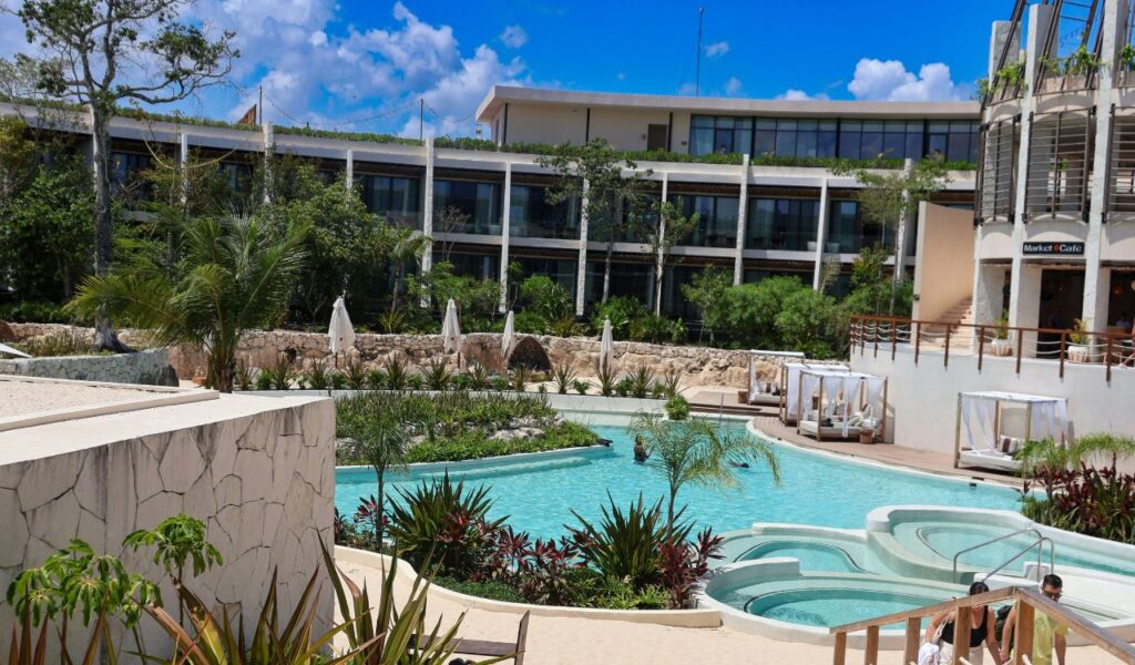 A tropical all-inclusive resort with a large pool, lush greenery, cabanas, and a modern hotel building under a sunny blue sky.