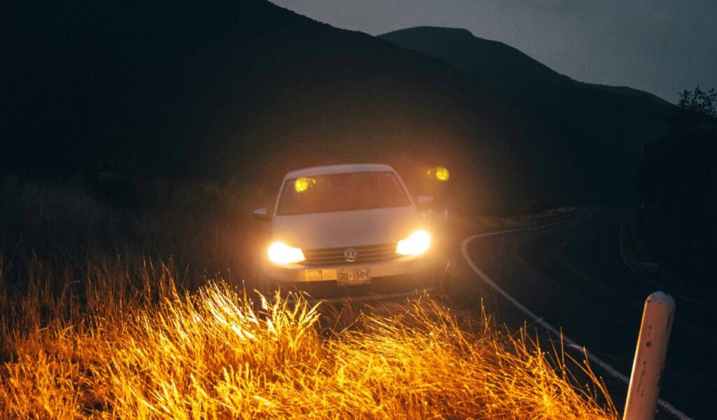 A car with headlights on driving along a dark rural road surrounded by hills and tall grass, illuminating the foreground in a nighttime setting.