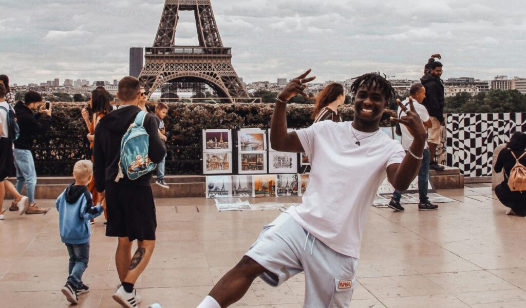 A smiling tourist poses playfully in front of the Eiffel Tower in Paris while other visitors take photos and enjoy the iconic landmark.