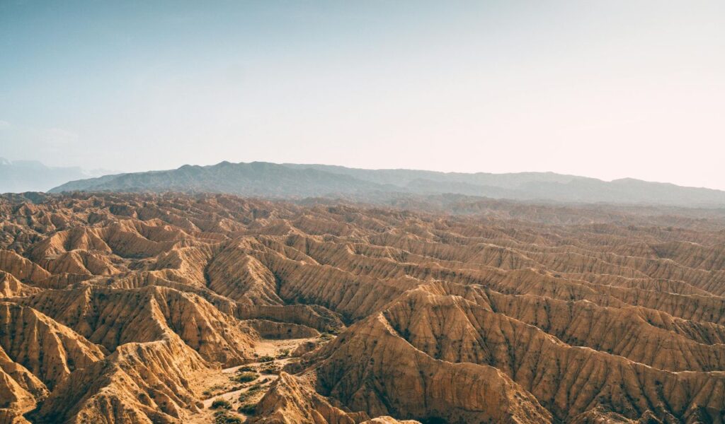 Vast badlands landscape with rugged ridges and dry valleys stretching toward distant hazy mountains under a pale sky.